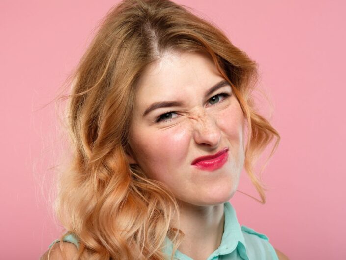 Egy fiatal nő közelről látható, fintorogva ráncolja az orrát és a homlokát, arckifejezése enyhe undort vagy elégedetlenséget sugall, rózsaszín háttér előtt.