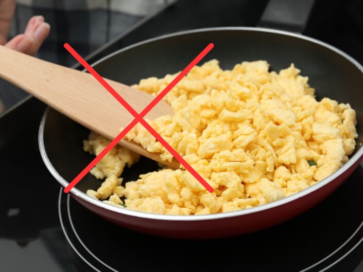 Serpenyőben készülő rántotta, amelyet fakanállal kevernek, a képen piros X jel mutatja, hogy ez a módszer nem ajánlott a tökéletes állag eléréséhez.