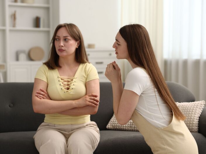 két hosszú barna hajú lány a kanapén beszélgetnek, az egyik magyaráz a másik sértett