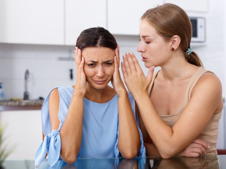 egy ujjatlan viselő lány csúnya dolgokat súg egy kék pólós lánynak, aki a fülére teszi a kezét