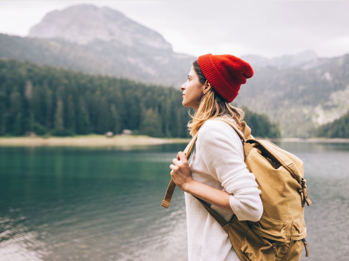 kiránduló sapkás hátizsákos nő áll egy tóparton körülötte hegyek