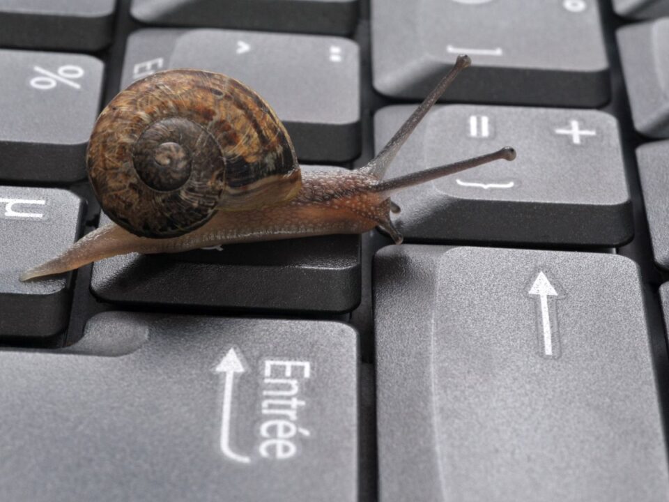Barna csiga kúszik egy fekete számítógép-billentyűzeten, az Enter és nyíl billentyűk közelében. A kép a lassú internetkapcsolatot jelképezi.