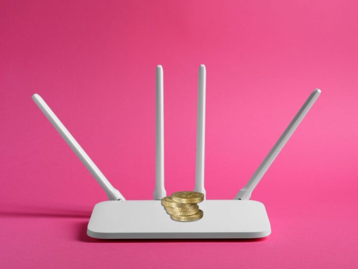 Fehér Wi-Fi router négy antennával, tetején néhány aranyszínű érme. Rózsaszín háttér előtt áll, a kép a netes „érmés trükkre” utal.