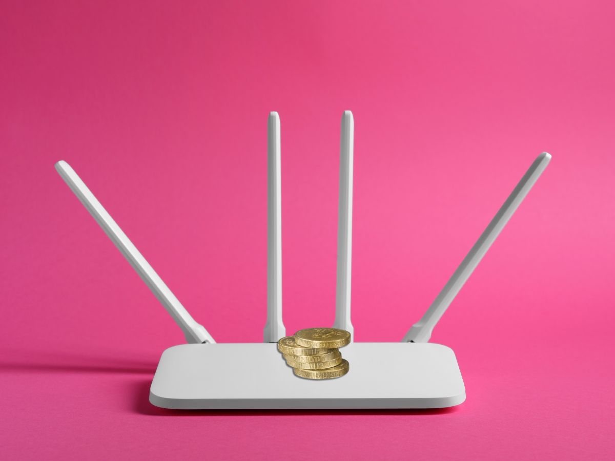 Fehér Wi-Fi router négy antennával, tetején néhány aranyszínű érme. Rózsaszín háttér előtt áll, a kép a netes „érmés trükkre” utal.