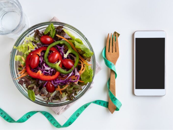 Színes zöldsaláta paradicsommal, paprikával és káposztával egy üvegtálban, mellette mérőszalag és telefon – a diéta és tudatos étkezés szimbóluma.