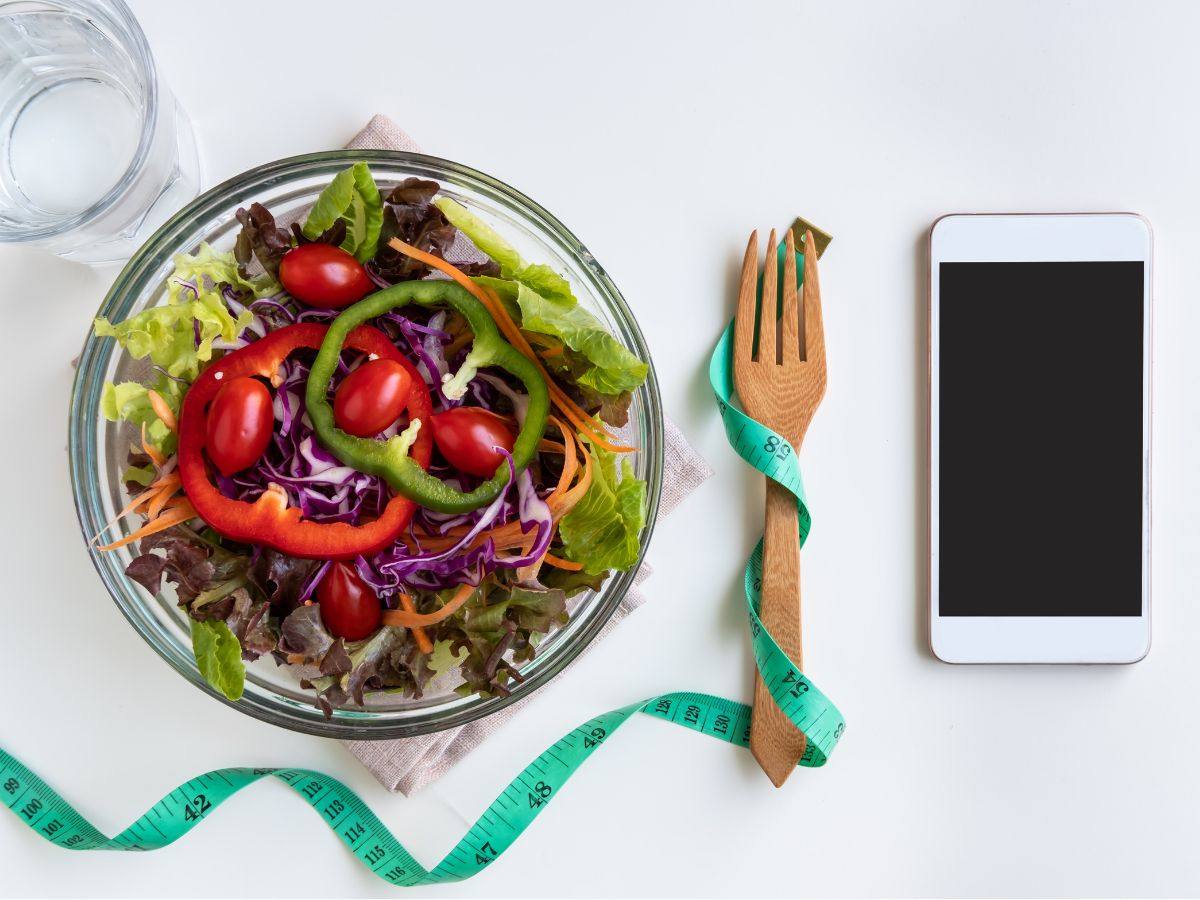 Színes zöldsaláta paradicsommal, paprikával és káposztával egy üvegtálban, mellette mérőszalag és telefon – a diéta és tudatos étkezés szimbóluma.