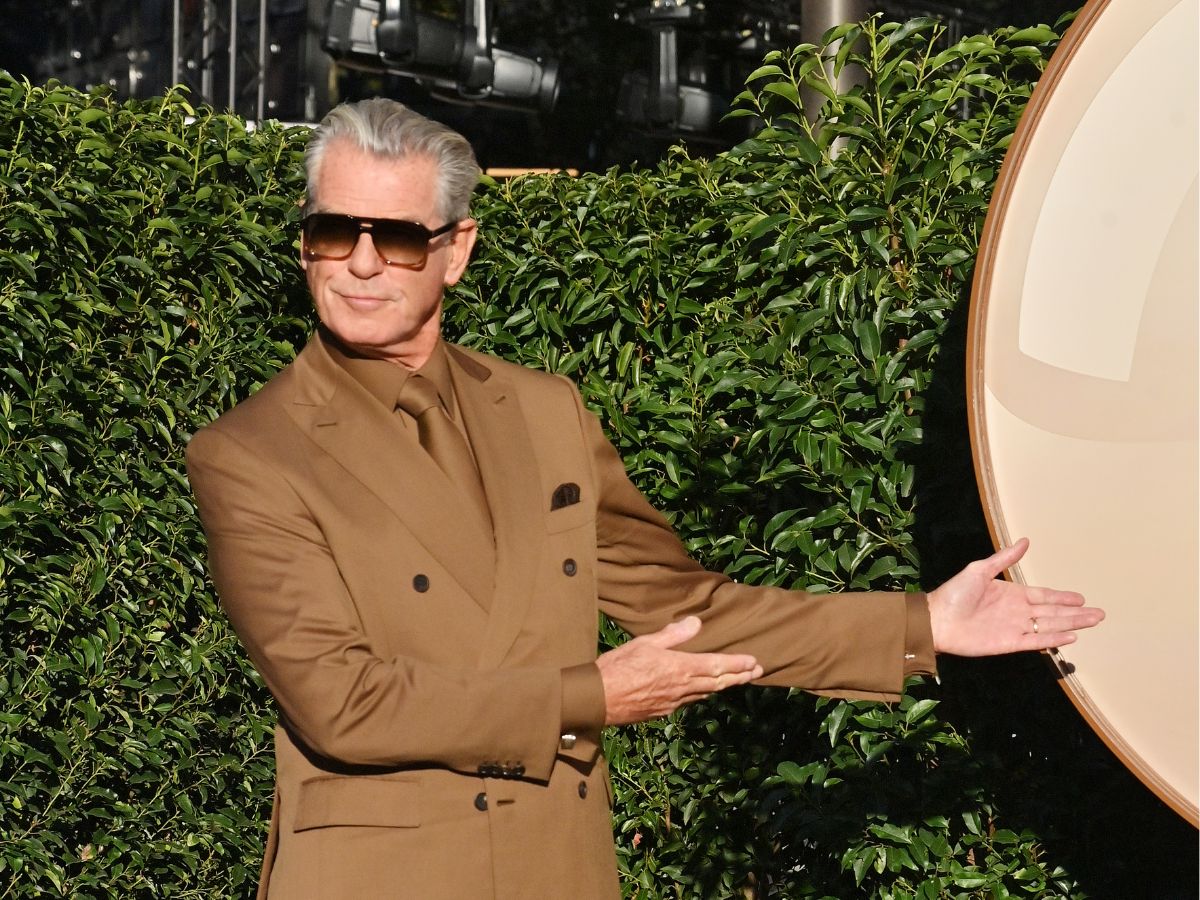 Pierce Brosnan napszemüvegben zöld növényfal előtt barna öltönyben mutat a kezeivel oldalra
