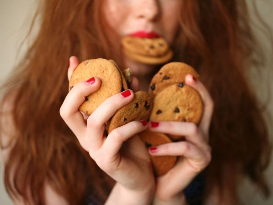 Hosszú, barna hajú nő kezében rengeteg csokis keksszel, elmosódva látszik, hogy a szájában is van