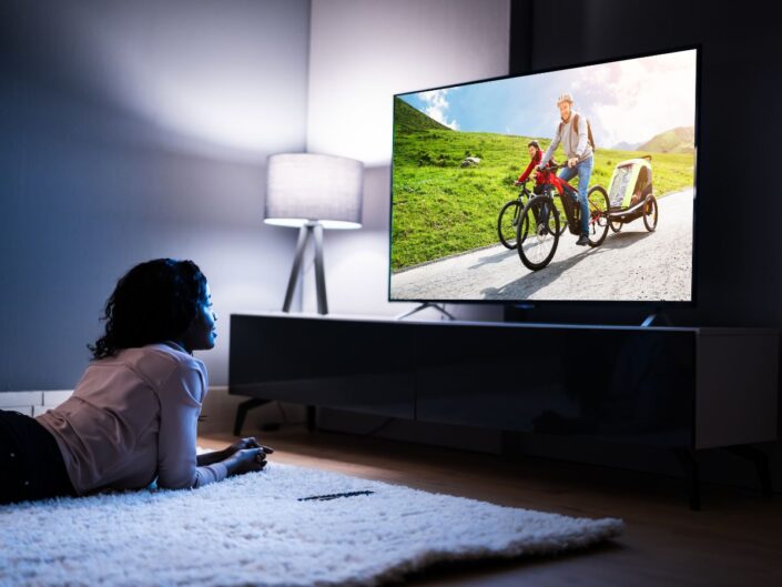 Egy nő sötét nappaliban hason fekve néz egy nagy képernyős okostévét, amelyen bicikliző család látható. A szoba hangulatos, a tévé erős fénnyel világítja meg az arcát.