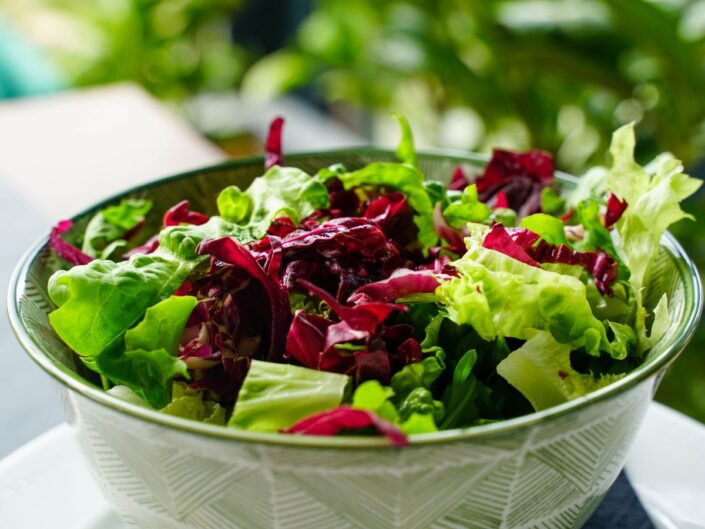 Egy tál friss, színes saláta közelről: zöld levelek, lila radicchio és ropogós darabok keverednek egy mintás tálban, világos, természetes fényben.