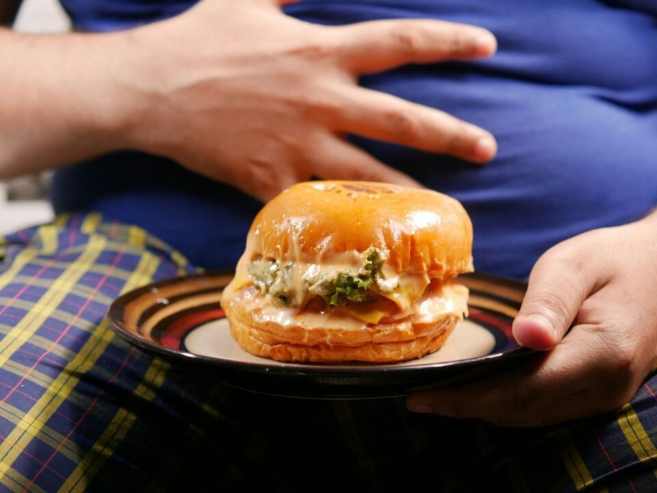 Férfi hasi zsírra teszi a kezét, ölében barna tányéron nagyméretű hamburger