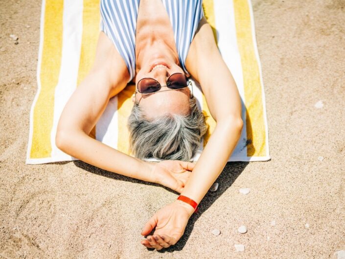 50 feletti, ősz hajú szép nő csíkos fürdőruhában fekszik sárga-fehér csíkos fürdőlepedőn homokos strandon napszemüvegben, kezét a feje fölé nyújtva