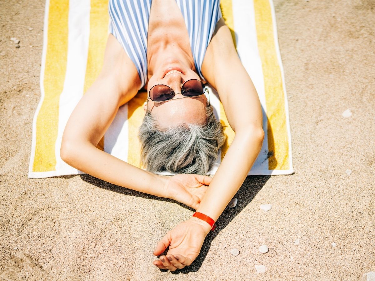 50 feletti, ősz hajú szép nő csíkos fürdőruhában fekszik sárga-fehér csíkos fürdőlepedőn homokos strandon napszemüvegben, kezét a feje fölé nyújtva