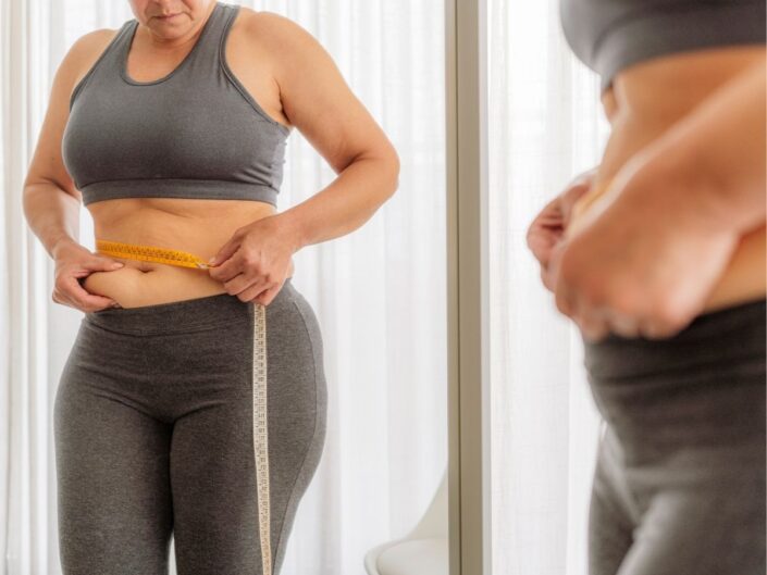 Nő szürke leggingszben és sportmelltartóban tükör előtt vizsgálja centivel a kezében a hasán lévő zsírt