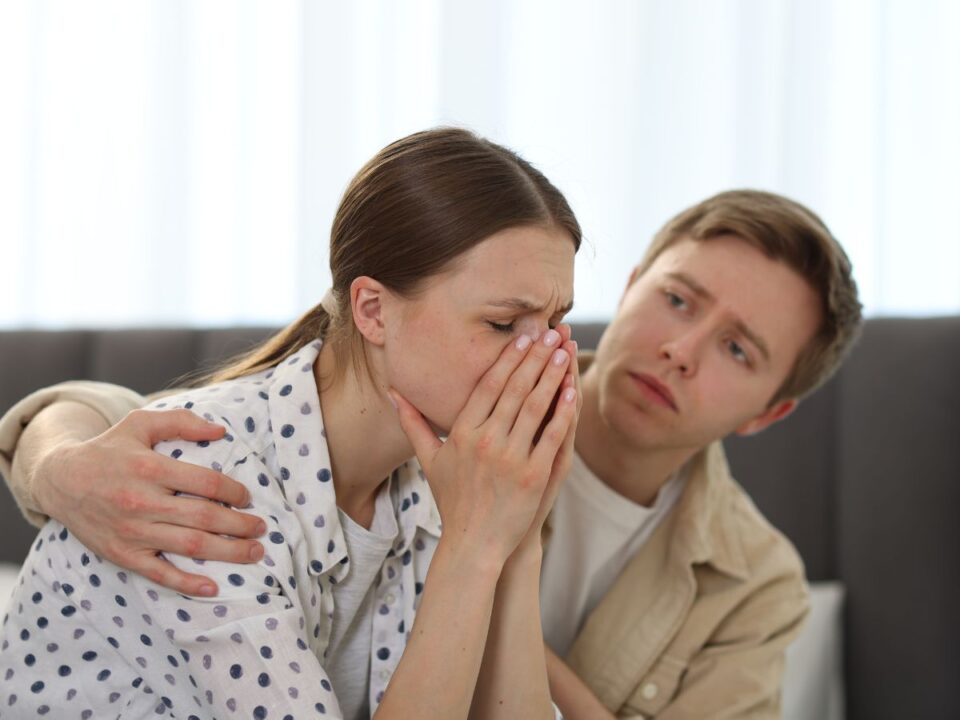 vita-kozbeni-siras - Nők Lapja Egy fiatal nő egy otthoni beszélgetés közben sír, kezével az arcát takarja. Mellette egy fiatal férfi aggódó arckifejezéssel átöleli és vigasztalni próbálja. A jelenet érzelmileg megterhelő helyzetet sugall.