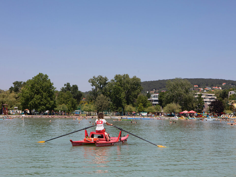 vízimentők Balaton copy - Nők Lapja kis piros csónakszerűségen evez egy vízimentő nő a Balatonon, a víz felől látjuk, háttérben a part, háttal van nekünk