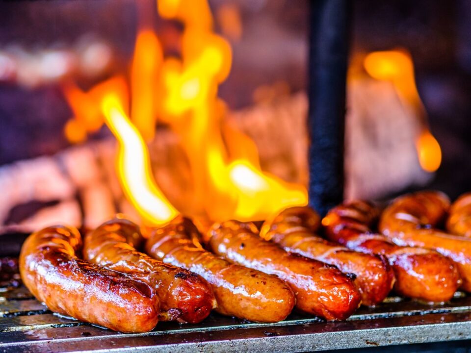 Grillen sülő kolbászok nyílt láng felett. A húsok barnulnak, a háttérben erős tűz látható.