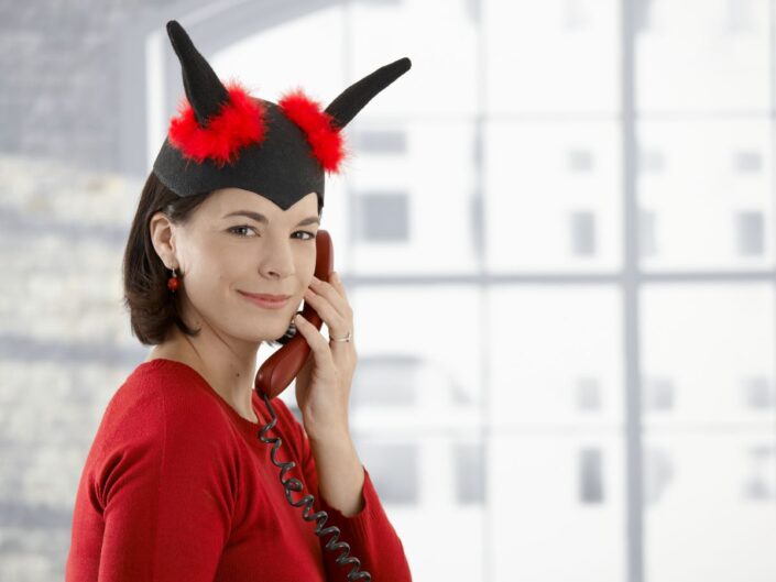 Egy vörös felsőt és fekete, szarvas fejdíszt viselő, mosolygó nő telefonál egy világos irodában.