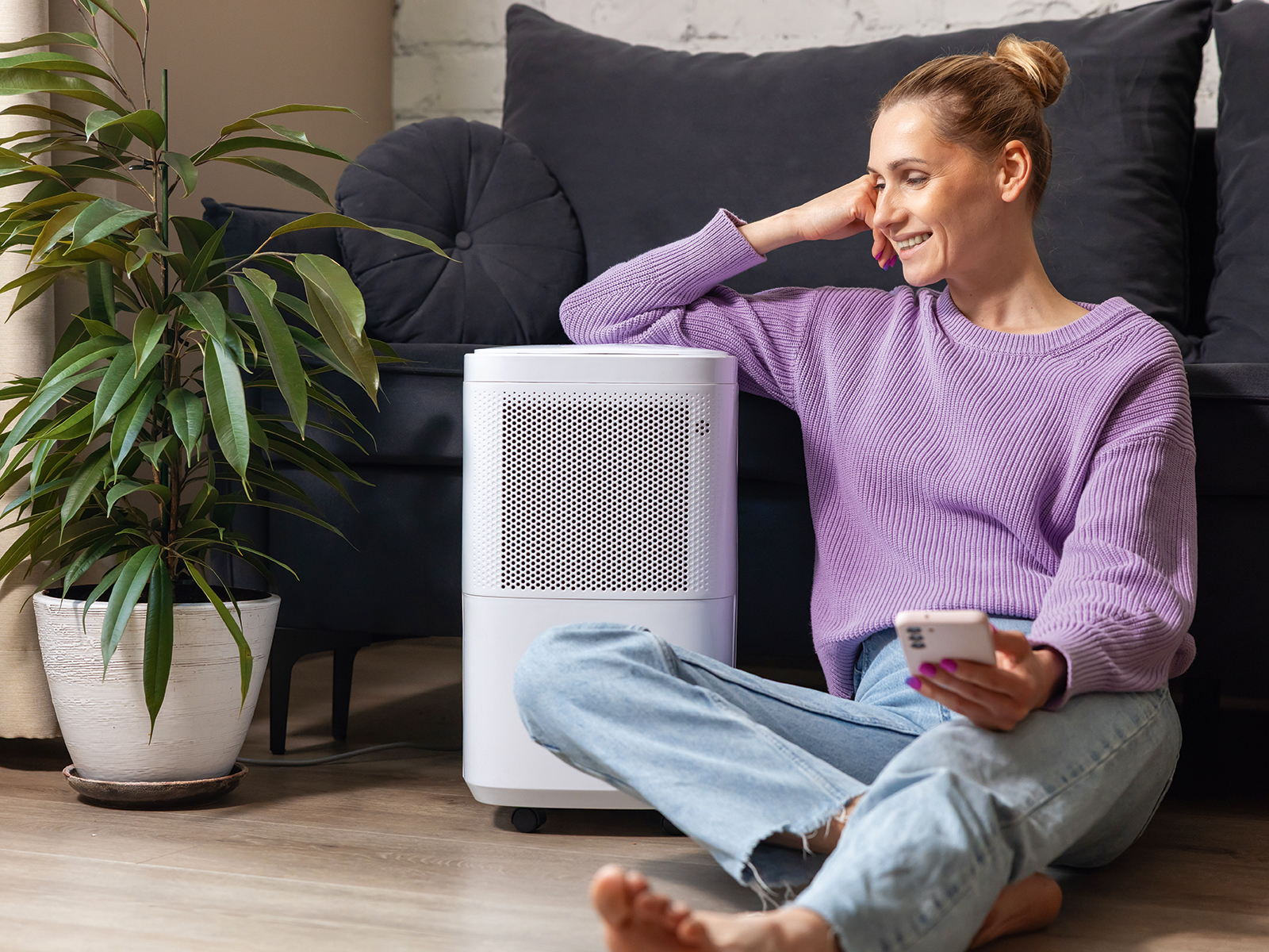 egy nő halványlila pulóverben és világoskék nadrágban ül egy nappalibana. földön egy sötétszürke kanapé előtt, jobb könyökét egy légtisztító készülékre helyezve, bal kezében mobiltelefon