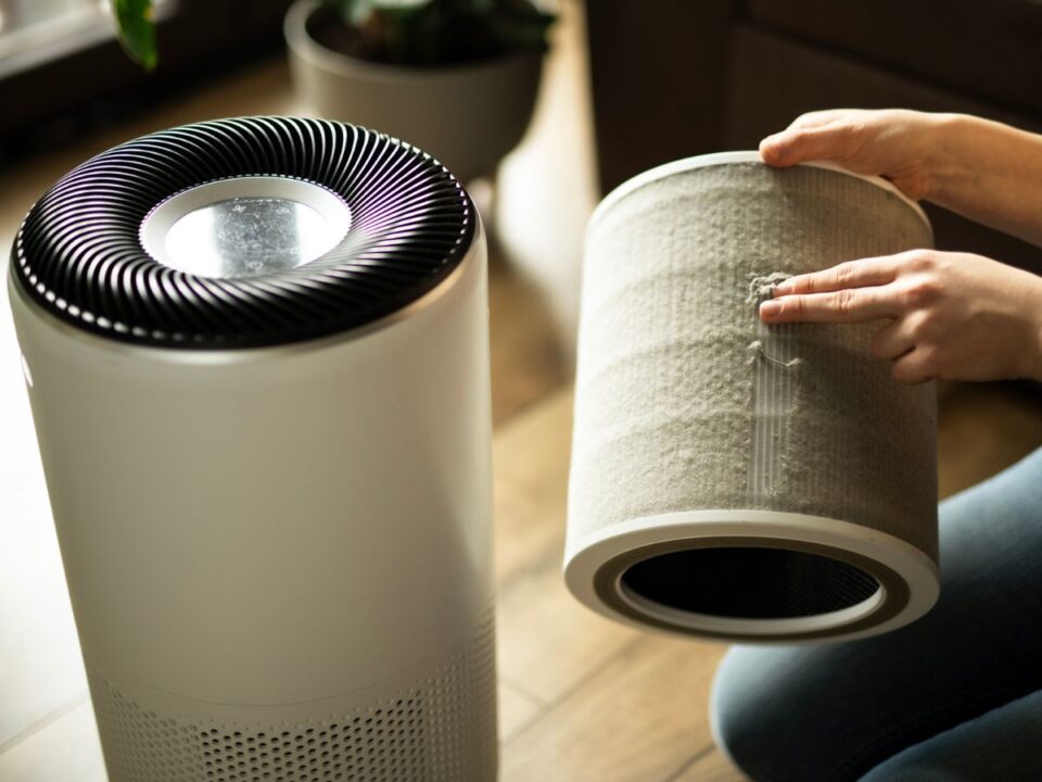 hepa-legszuro-allergia - Nők Lapja Egy kéz egy henger alakú, porral szennyezett légszűrőbetétet tart egy fehér, otthoni légtisztító készülék mellett, a szűrő cseréjét vagy tisztítását szemléltetve.