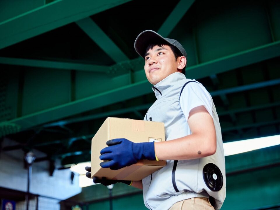 japan-ventilatoros-kabat - Nők Lapja Egy csomagot szállító futár világosszürke ventilátoros mellényt visel. Az oldalába épített ventilátor jól látható, miközben dobozt tart a kezében.