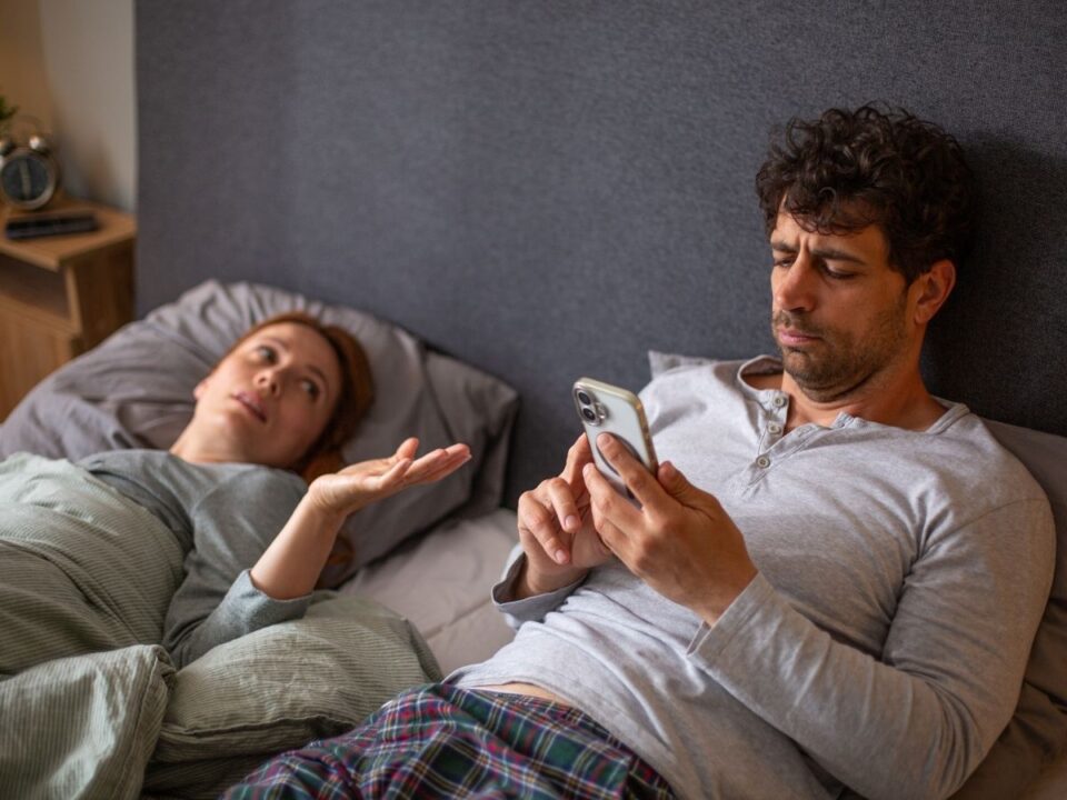 megcsalas-jelei-telefonozo-ferfi - Nők Lapja Ágyban fekvő férfi és nő, a férfi telefonozik, miközben a nő beszél hozzá
