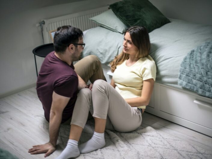 Fiatal pár ágy mellett a földön ülve beszélget