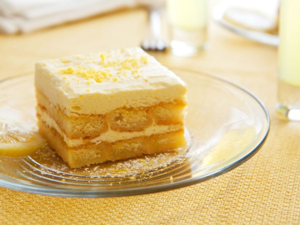 sutes-nelkuli-citromos-alom-recept - Nők Lapja Négyzet alakú szelet sütés nélküli citromos sütemény üvegtányéron. A krémrétegek között babapiskóta látszik, a tetejét citromhéj és porcukor díszíti.