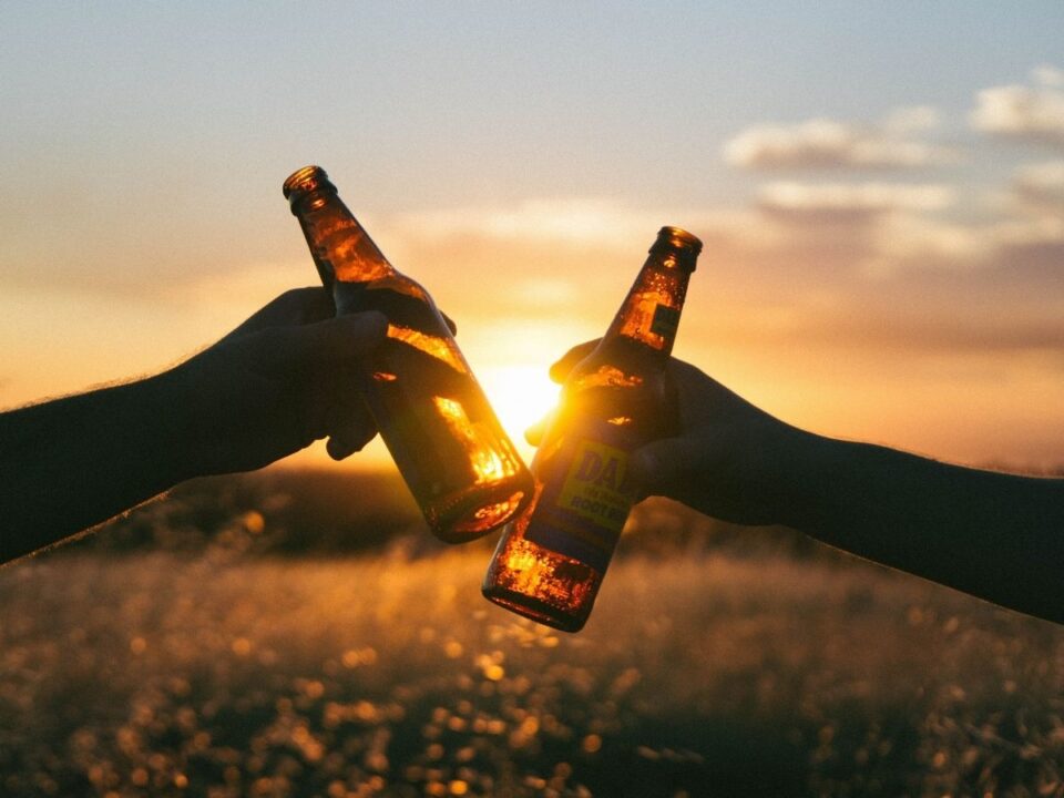 alkoholmentes-sor-gyerek-kockazat - Nők Lapja Két ember barna üvegekkel koccint naplementében; a kezek és az üvegek sötét sziluettje előtt aranyszínben ragyog a táj.
