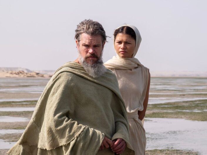 Jelenetkép Christopher Nolan Odüsszeia filmjéből, a képen Matt Damon (Odüsszeusz) és Zendaya (Pallasz Athéné) állnak homokszínű és fehér ruhákhban, mögöttük a ruháik színéhez hasonló táj, vizej talaj