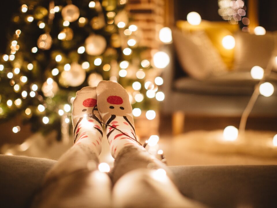 december-24-munkaszuneti-nap - Nők Lapja Karácsonyi mintás zoknit viselő lábak pihennek egy kanapén, körülöttük fényfüzér világít, a háttérben feldíszített karácsonyfa és kandalló látszik.
