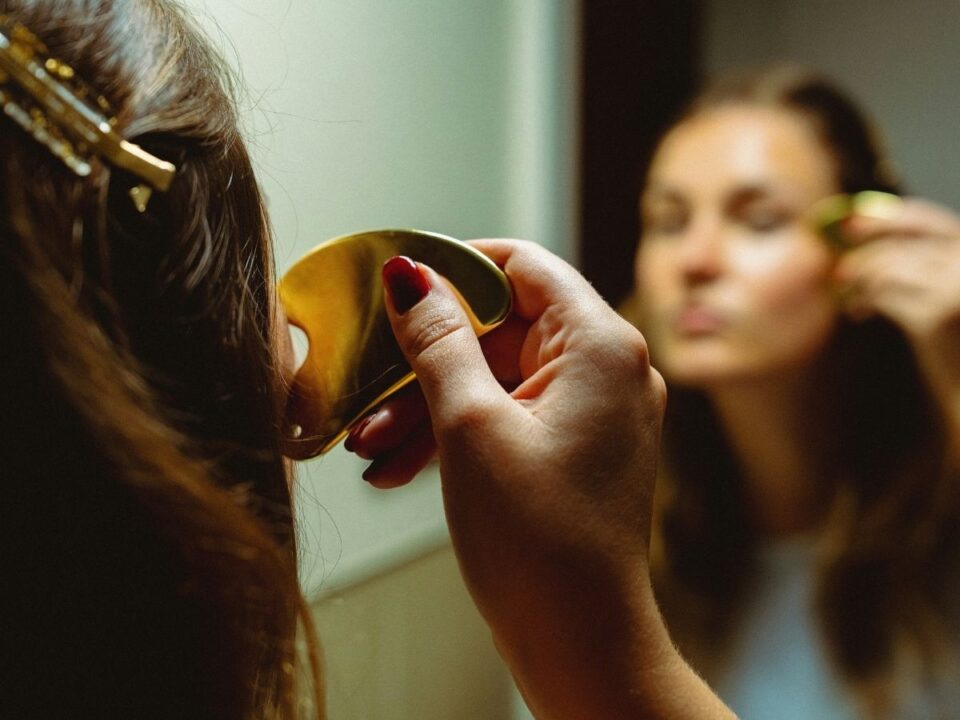 gua-sha-arcmasszazs - Nők Lapja Nő tükör előtt arany gua sha kővel masszírozza az arcát háttal a kamerának