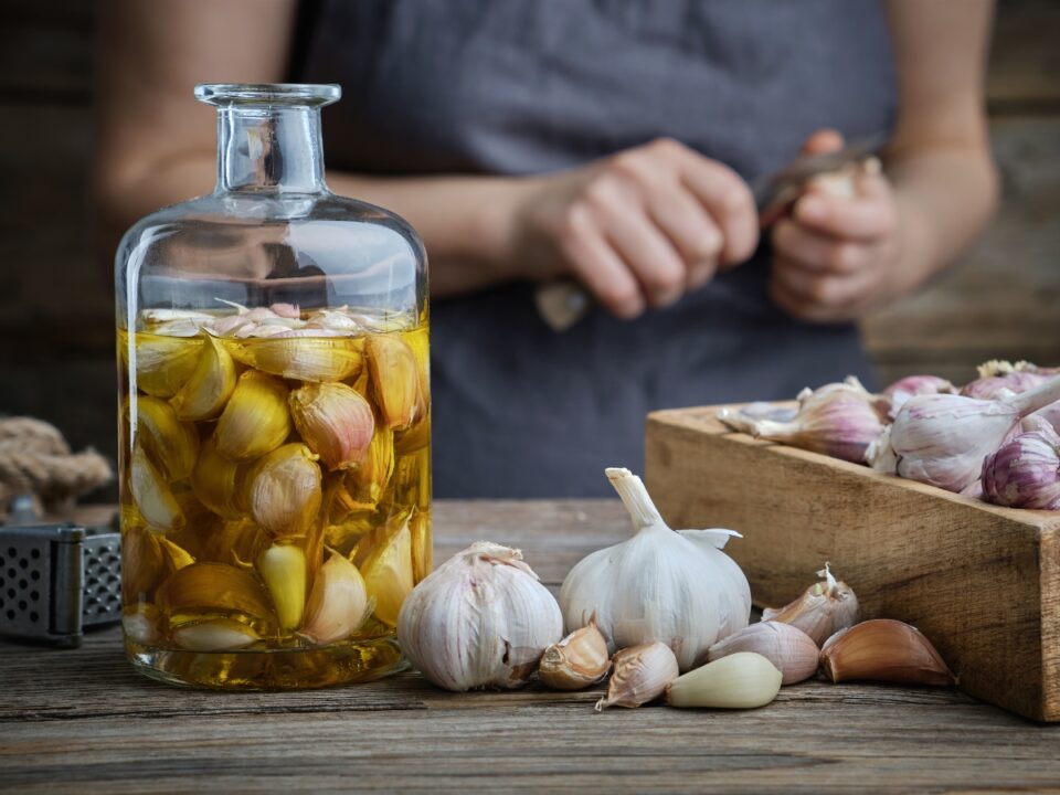 ikaria-fokhagyma-hosszu-elet - Nők Lapja Olívaolajjal és egész gerezdekkel töltött üveg áll a faasztalon, mellette fokhagymafejek és gerezdek; a háttérben valaki fokhagymát pucol.