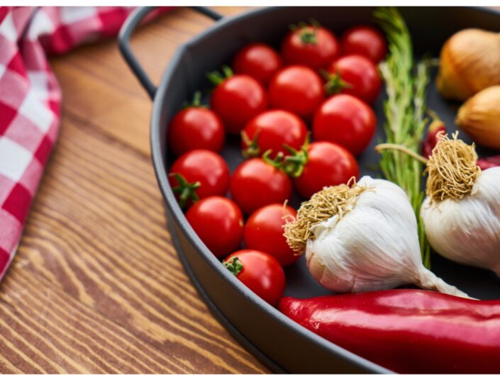 Fekete sütőtálban piros koktélparadicsomok, két egész fokhagyma, rozmaring és hosszú piros paprika látható egy faasztalon.