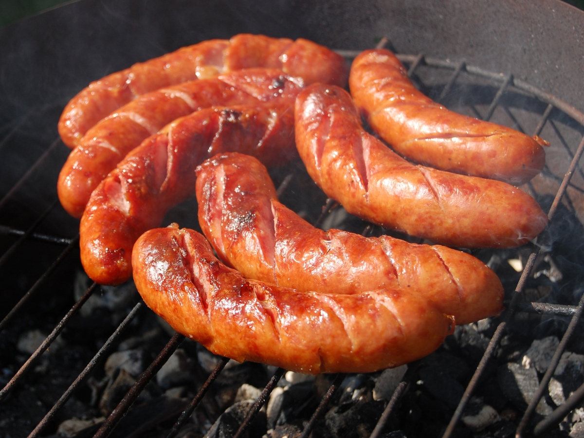 Fényesre sült, bevagdalt kolbászok sorakoznak egy szabadtéri, faszenes grillsütő rácsán, miközben vékony füst száll fel körülöttük.