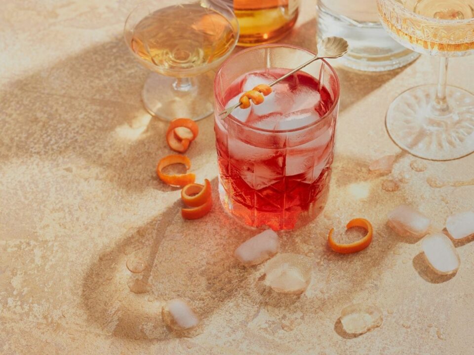 negroni-koktel-elso-randi - Nők Lapja Negroni koktél bézs asztalon, narancshéj, jégkockák és más koktélos poharak körülötte