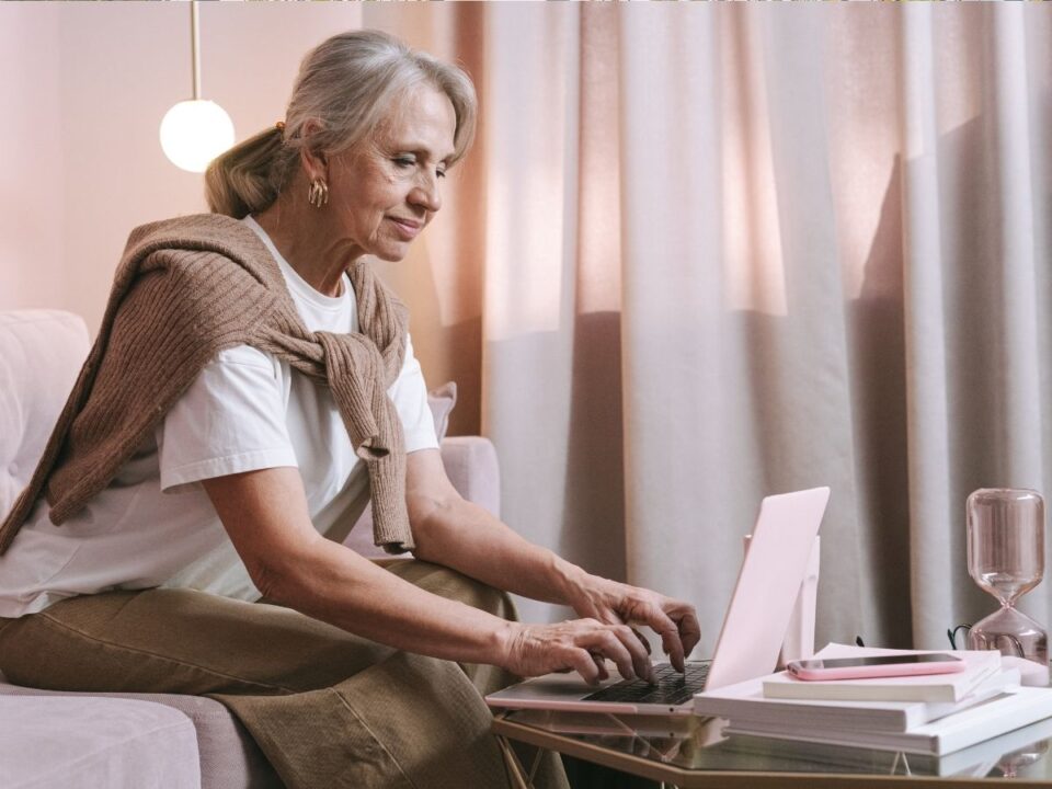 idősebb ősz hajú nyakába kötött barna pulóverrel gépel a laptopján