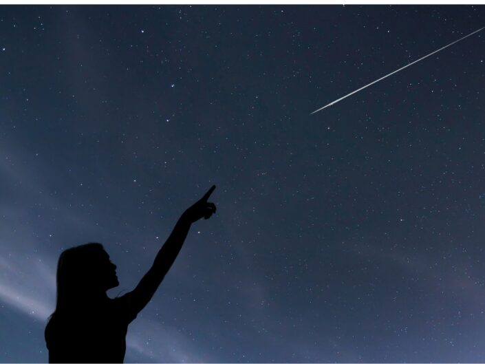 Egy nő sötét sziluettje az ég felé mutat, miközben a csillagokkal teleszórt éjszakai égbolton hosszú, fényes meteorcsík húzódik.