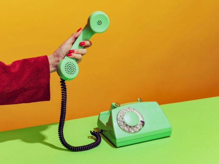 Neon narancssárga és zöld háttér, piros kabátujjas női kéz kivizöld telefonkagylót tart
