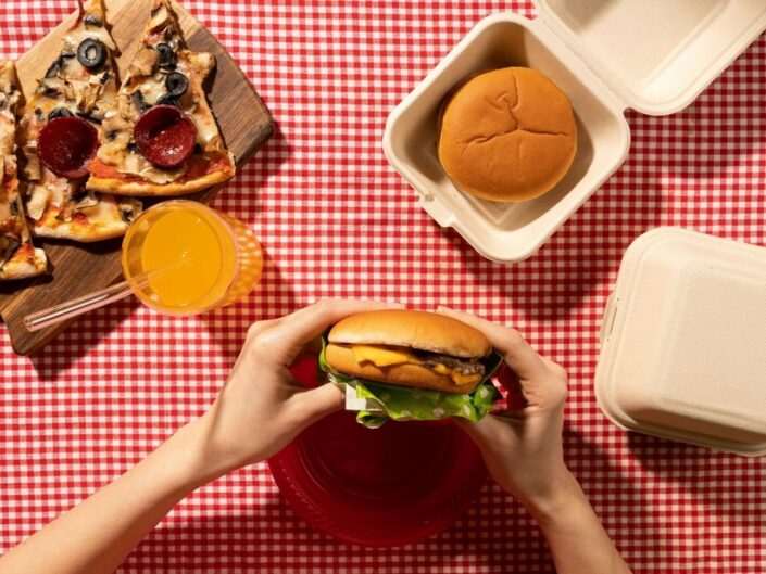 Nő gyorséttermi hamburgert tart a kezében piros kockás terítős asztalnál, mellette narancsos üdítő, pizza