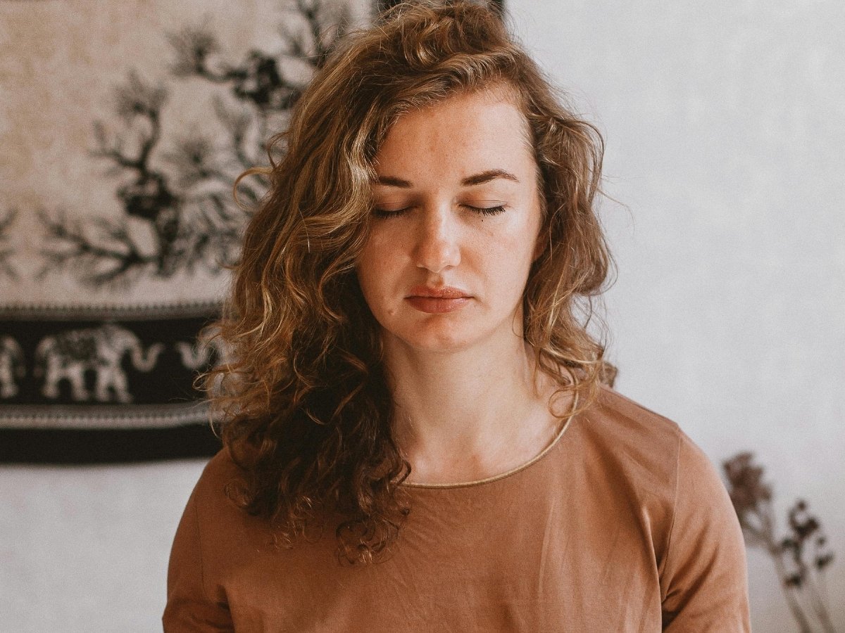 Göndör, barna hajú nő ül lehunyt szemmel, barna felsőben. Mögötte világos fal és egy bekeretezett, növényi mintás fekete-fehér kép látható.
