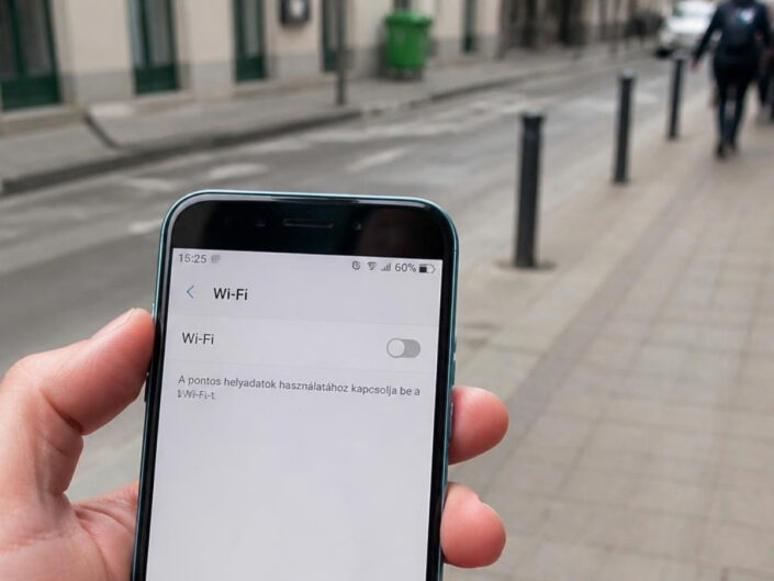 Egy kéz telefont tart az utcán. A kijelzőn a Wi-Fi-beállítások láthatók, a kapcsoló kikapcsolt állásban van.