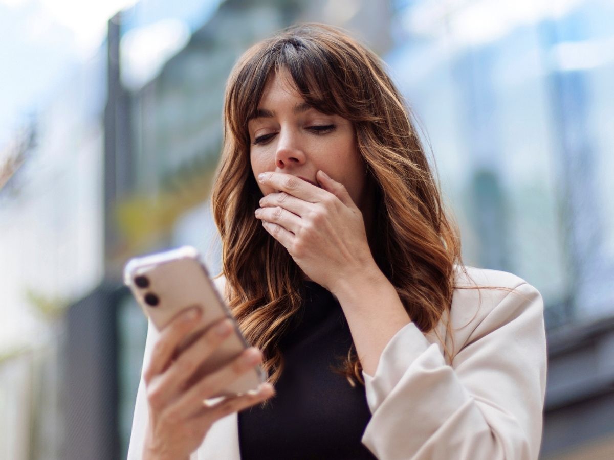 Meglepett nő kissé alulról fotózva száját bal kezével eltakarva nézi a jobb kezében trtott telefonja képernyőjét
