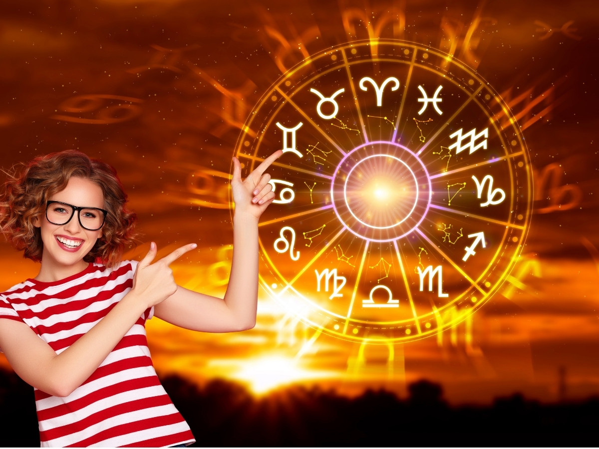Mosolygó, szemüveges nő piros-fehér csíkos felsőben egy fénylő, aranyszínű zodiákuskörre mutat, narancssárga, naplementés háttér előtt.