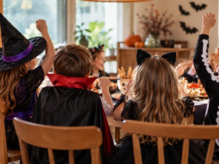 Halloweenjelmezbe öltözött gyerekek ülnek egy ünnepi finomságokkal megrakott asztal körül. Hátulról látszanak, többen örömükben felemelik a kezüket.