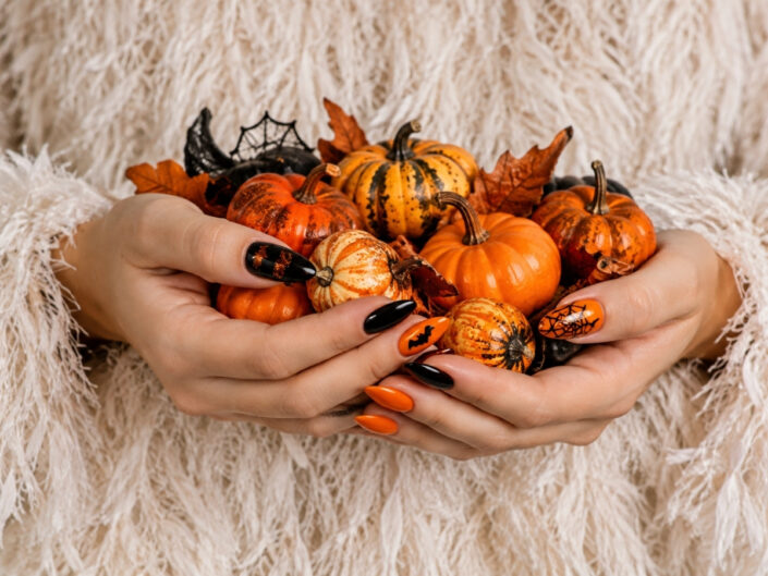 Nő fehér, tollszerű szőrös felsőben apró halloween tököket tart a kezében, hangsúlyos narancs-fekete halloween manikűrrel