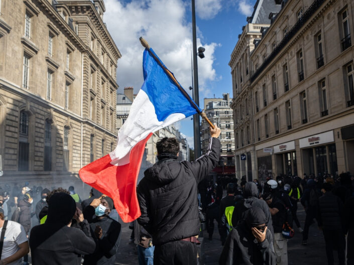 Franciaországi tüntetésen fekete ruhás tüntetők vonulnak, középen férfi francia nemzeti lobogót emel magasba 2026. október 17-én