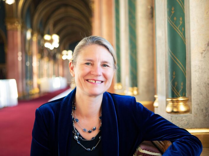 Tanja Gonggrijp, az Európa Tanács főtitkárhelyettese sötétkék bársonykabátban, mosolyogva ül egy díszes, oszlopos folyosón.