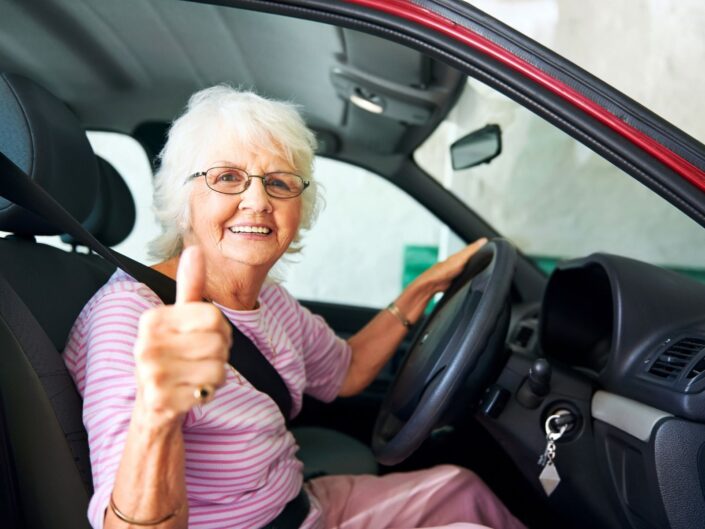 Mosolygó idős nő ül egy autó volánjánál, egyik kezével a kormányt fogja, a másikkal felfelé mutatja a hüvelykujját.