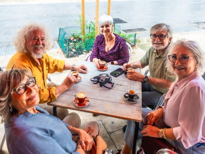 Öt idősebb ember, három nő és két férfi mosolyog egy kávézó teraszán az asztal körül. Előttük kávéscsészék, az asztalon telefon és fényképezőgép látható.